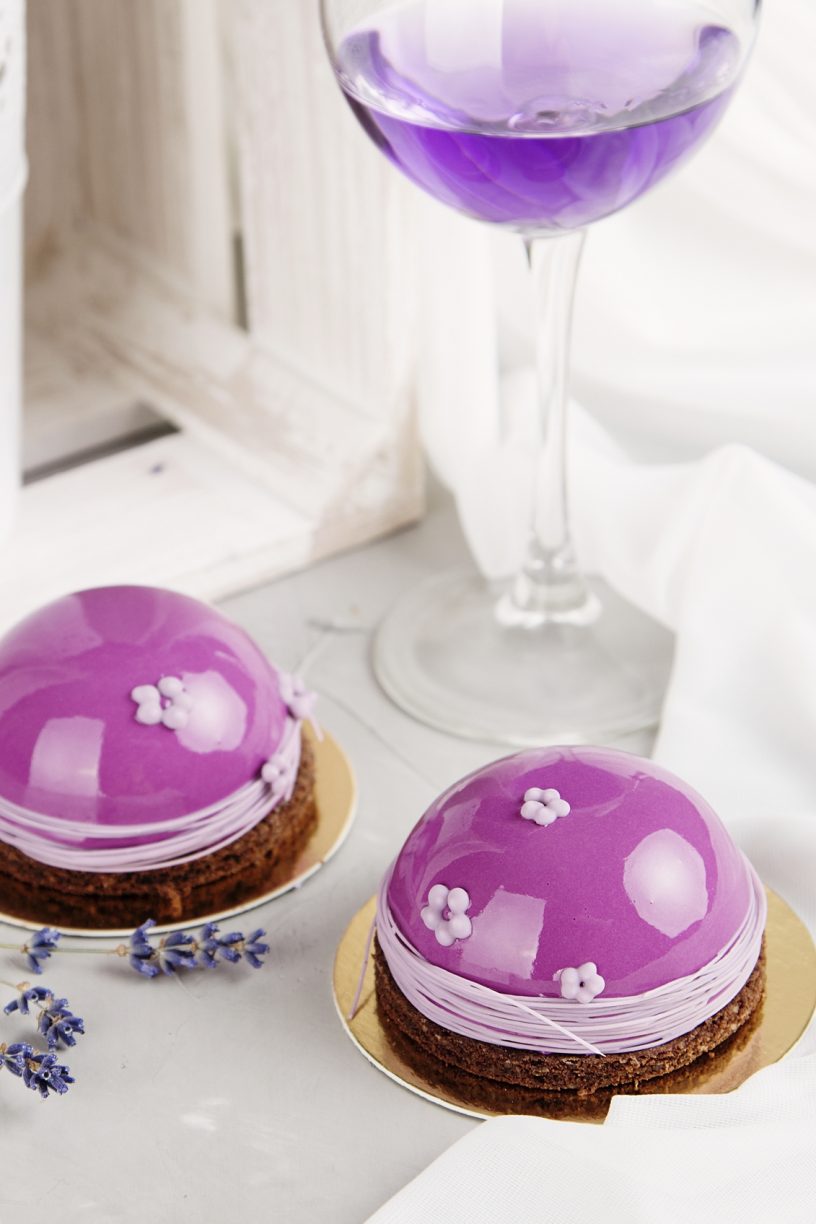 Romantic french pastries with pink mirror glaze cover on pastel background with wine glass and lavender flowers. Romantic holiday concept. Shallow focus